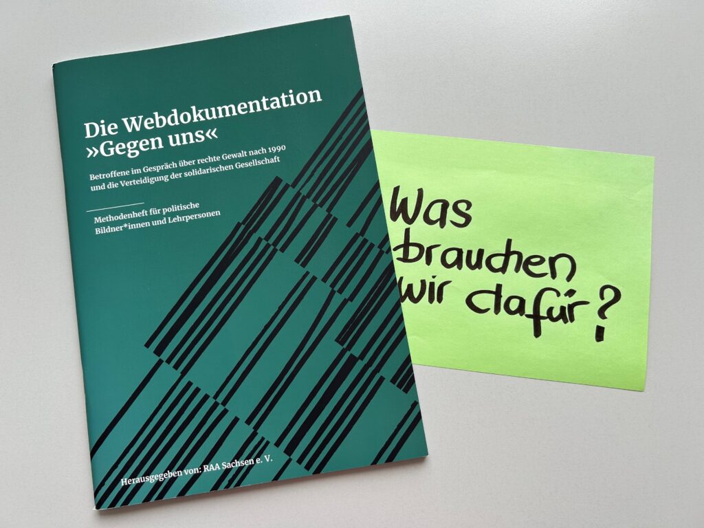 Foto der Broschüre "Die Webdokumentation 'Gegen uns'", daneben liegt ein großes Post-It, darauf die Frage geschrieben: "Was brauchen wir dafür?"